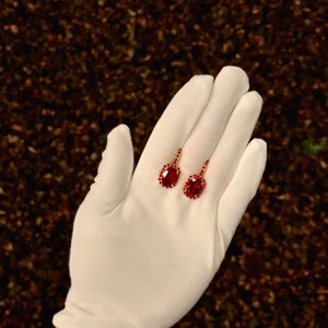 May include: A pair of red gemstone earrings with a gold setting. The earrings are oval shaped and have a halo of smaller gemstones around the center stone.