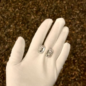 May include: A pair of rectangular-cut diamond earrings with a halo of smaller diamonds, displayed on a white gloved hand. The earrings have a silver setting and are set against a textured brown background.