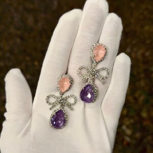 May include: A pair of dangle earrings with a pink and purple teardrop-shaped stone and a silver bow. The earrings are on a white surface.