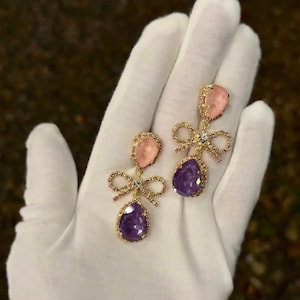 Op de afbeelding: Een paar gouden oorbellen met een roze en paarse druppelvormige steen en een strikdetail.