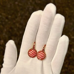 May include: A pair of gold earrings with red and white striped round charms. The charms are surrounded by a circle of small red rhinestones.