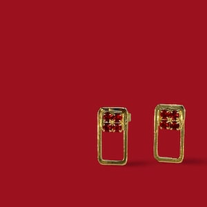 May include: A pair of gold earrings with a red stone design. The earrings are rectangular in shape and have a simple, modern style.