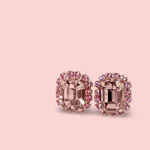 May include: A pair of pink stud earrings. Each earring has a square, light pink central stone, encircled by smaller pink and rose-coloured crystals. The earrings are set against a soft pink background.