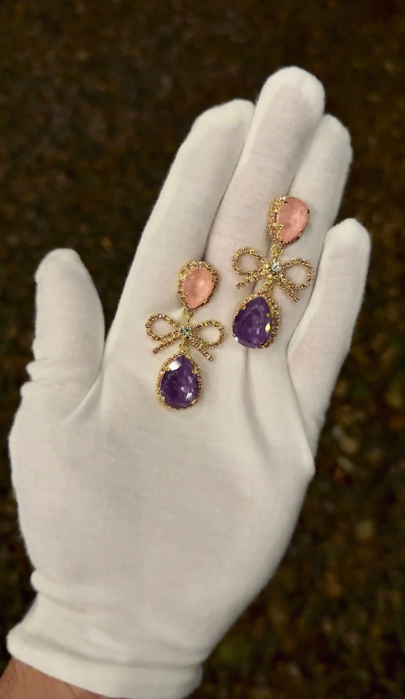 Op de afbeelding: Een paar gouden oorbellen met een roze en paarse druppelvormige steen en een strikdetail.