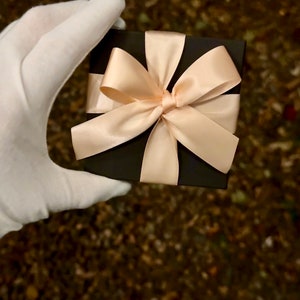 Op de afbeelding: Een zwarte cadeaudoos met een grote, cr&egrave;mekleurige satijnen strik. De doos wordt vastgehouden door een hand met een handschoen.