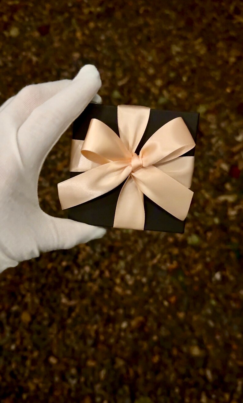 Op de afbeelding: Een zwarte cadeaudoos met een grote, cr&egrave;mekleurige satijnen strik. De doos wordt vastgehouden door een hand met een handschoen.