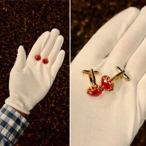 May include: A pair of gold cufflinks with red jeweled centers. The cufflinks are on a white surface.