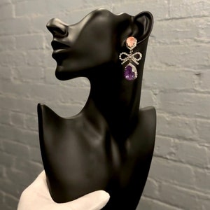 May include: A pair of dangle earrings with a pink and purple stone, a silver bow, and clear rhinestones.