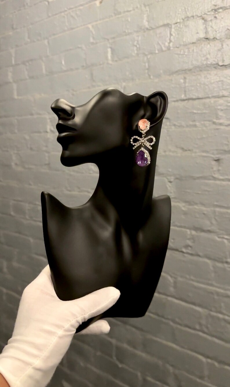 May include: A pair of dangle earrings with a pink and purple stone, a silver bow, and clear rhinestones.