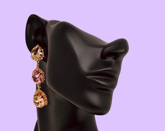 May include: A black mannequin wearing a pair of gold dangle earrings with pink and clear teardrop-shaped crystals.