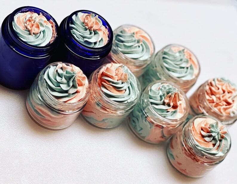 May include: A collection of small glass jars filled with a swirl of light pink and light green whipped cream. The jars are arranged in a row on a white surface.