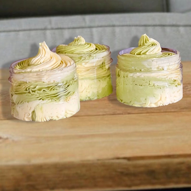 May include: Three glass jars filled with a green and white whipped body butter. The jars are on a wooden surface.