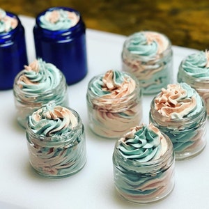 May include: Eight small glass jars filled with a swirl of light pink and light blue whipped cream. The jars are arranged on a white surface.