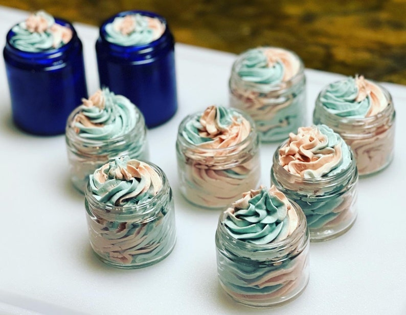 May include: Eight small glass jars filled with a swirl of light pink and light blue whipped cream. The jars are arranged on a white surface.