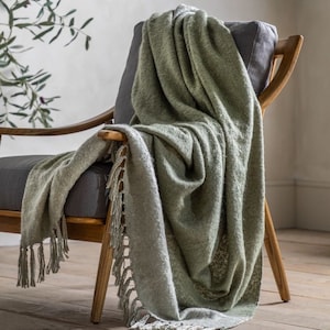 May include: A soft, green throw blanket with fringe detailing draped over a wooden chair. The blanket has a subtle texture and a light, airy feel.