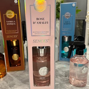 May include: A selection of SENCES home fragrance products. The image features a Rose D'Amalfi boutique diffuser in a pink box, with a clear bottle inside. Other boxes include Vanilla Antique and Aqua Marine. A pink spray bottle is also visible.