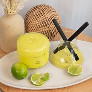 May include: Two yellow glass jars with black lids and black sticks inside. The jars are labelled "Serenity Lime Splash". The jars are sitting on a white oval plate with lime slices on the plate.