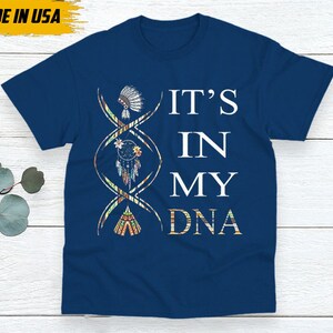 May include: Navy blue t-shirt with the text "IT'S IN MY DNA" in white. The design includes a DNA strand with Native American-inspired illustrations of a headdress, dreamcatcher, and teepee. The shirt is labeled "MADE IN USA".