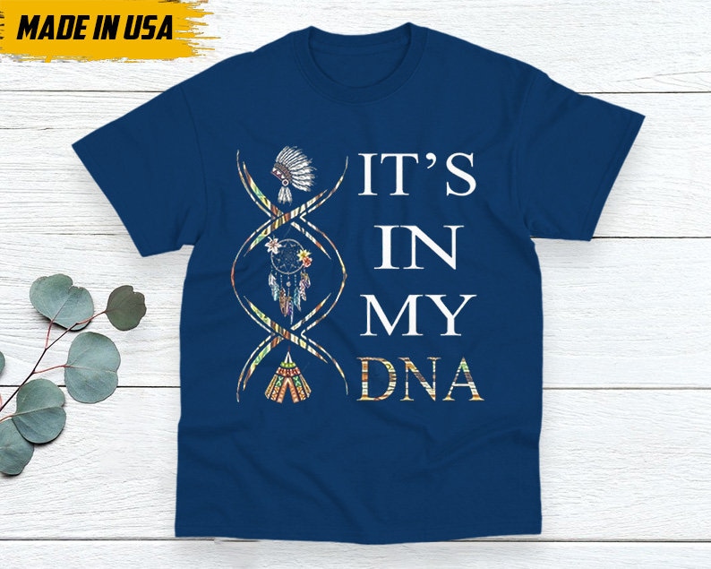 May include: Navy blue t-shirt with the text "IT'S IN MY DNA" in white. The design includes a DNA strand with Native American-inspired illustrations of a headdress, dreamcatcher, and teepee. The shirt is labeled "MADE IN USA".