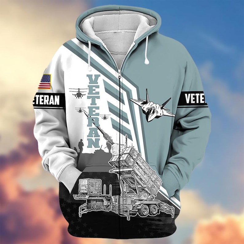 Pode incluir: Um casaco com capuz e z&iacute;per com tema militar. O design apresenta um jato, helic&oacute;pteros e um lan&ccedil;ador de m&iacute;sseis. A palavra "VETERAN" &eacute; impressa verticalmente na frente, com "VETE" na manga. Uma bandeira americana est&aacute; na manga esquerda.