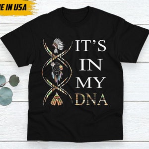 May include: Black t-shirt with the text "IT'S IN MY DNA" in white. The design includes a DNA strand with Native American-inspired illustrations of a headdress, dreamcatcher, and teepee. The words "MADE IN USA" are in a yellow box.