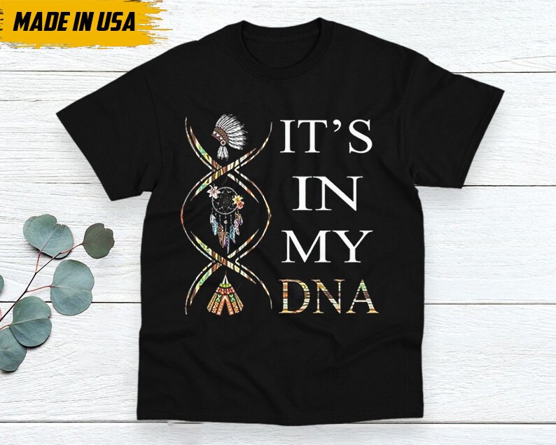 May include: Black t-shirt with the text "IT'S IN MY DNA" in white. The design includes a DNA strand with Native American-inspired illustrations of a headdress, dreamcatcher, and teepee. The words "MADE IN USA" are in a yellow box.