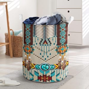 May include: A cylindrical laundry hamper with a Native American-inspired design. The hamper features a beige background with a pattern of turquoise, brown, and yellow geometric shapes and feather accents. White handles are attached to the top.