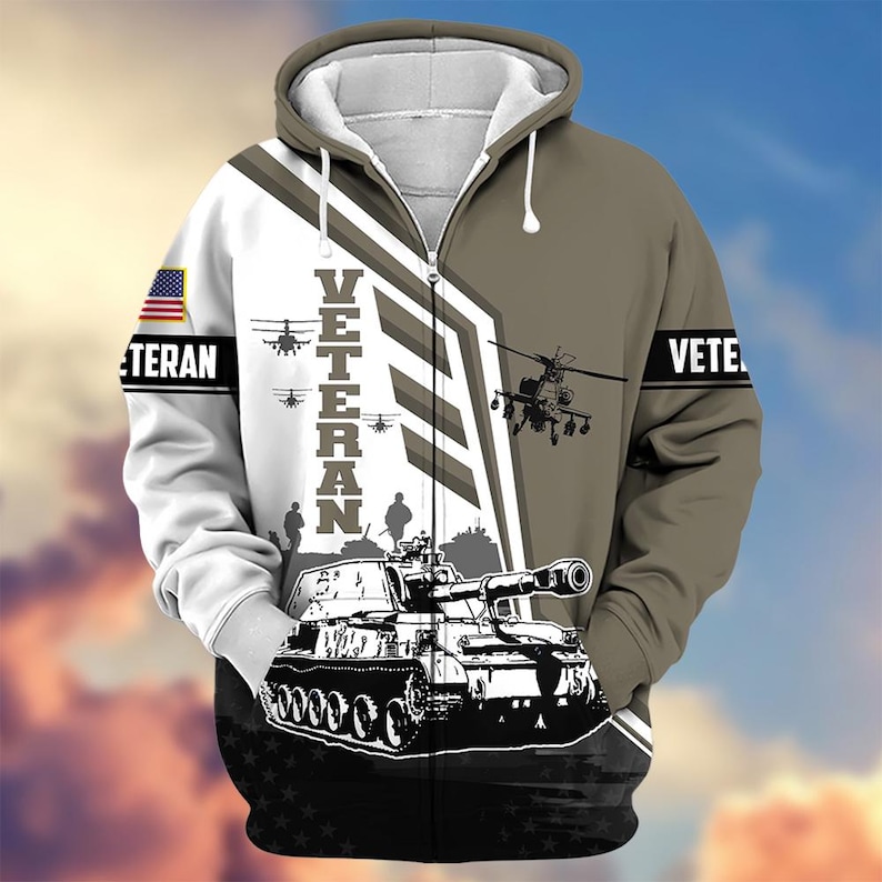 Pode incluir: Um casaco com capuz e z&iacute;per com tema militar. O design apresenta um tanque, helic&oacute;pteros e a palavra "VETERAN" em letras grandes. A bandeira americana est&aacute; na manga esquerda e a palavra "VETE" na manga direita.