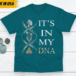 May include: Teal t-shirt with the text "IT'S IN MY DNA" in white. The design includes a DNA strand with Native American-inspired illustrations. The shirt has a tag that says "MADE IN USA".