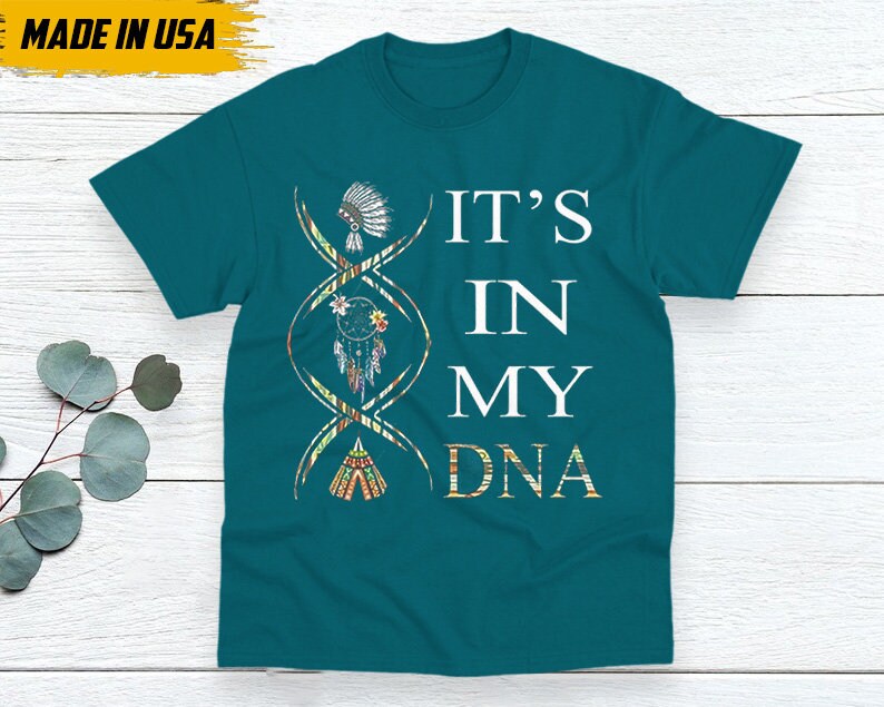 May include: Teal t-shirt with the text "IT'S IN MY DNA" in white. The design includes a DNA strand with Native American-inspired illustrations. The shirt has a tag that says "MADE IN USA".