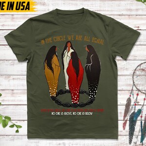 Puede incluir: Camiseta verde oliva con el texto "IN THE CIRCLE WE ARE ALL EQUAL" y "NO ONE IS ABOVE, NO ONE IS BELOW." La camiseta presenta una ilustraci&oacute;n de tres figuras en marr&oacute;n, blanco y rojo, de pie en un c&iacute;rculo. "MADE IN USA" est&aacute; impreso en la parte superior.