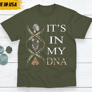 May include: Olive green t-shirt with the text "IT'S IN MY DNA" in white. A DNA strand graphic features Native American-inspired illustrations. The shirt has a crew neck and short sleeves. The words "MADE IN USA" are in a yellow box.