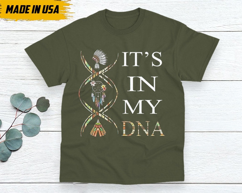 May include: Olive green t-shirt with the text "IT'S IN MY DNA" in white. A DNA strand graphic features Native American-inspired illustrations. The shirt has a crew neck and short sleeves. The words "MADE IN USA" are in a yellow box.