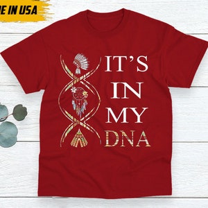 May include: Red t-shirt with the text "IT'S IN MY DNA" in white. The design includes a DNA strand with Native American-themed illustrations. The shirt also has a "MADE IN USA" label.