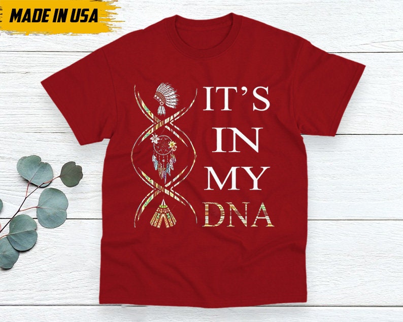 May include: Red t-shirt with the text "IT'S IN MY DNA" in white. The design includes a DNA strand with Native American-themed illustrations. The shirt also has a "MADE IN USA" label.