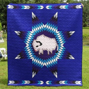 May include: A blue quilt with a central star design featuring a white bison. The star is composed of blue, white, and black geometric shapes. The quilt has a border with arrow-like patterns, creating a Native American-inspired design.