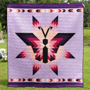May include: A large, lavender quilt featuring a geometric butterfly design in shades of black, pink, and purple. The butterfly is centered on the quilt, with a border of repeating arrow shapes in various colors. The quilt is displayed outdoors on a grassy lawn.