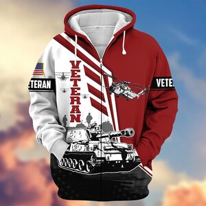 Pode incluir: Casaco com capuz e fecho de correr completo com um design patri&oacute;tico. O casaco apresenta um esquema de cores vermelho e branco com um gr&aacute;fico a preto e branco de um tanque e helic&oacute;pteros. A palavra "VETERAN" &eacute; impressa verticalmente na frente.