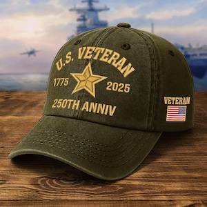 May include: Olive green baseball cap with gold embroidered text and star design. The text reads "U.S. VETERAN 1775 2025 250TH ANNIV" and "VETERAN" with an American flag patch. The cap is on a wooden surface.