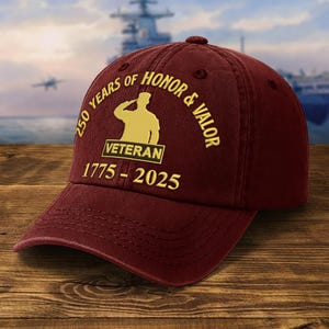 May include: A maroon baseball cap with gold text and a saluting soldier silhouette. The text reads "250 YEARS OF HONOR & VALOR" and "VETERAN 1775-2025". The cap is made of a soft material and has a curved brim. The background shows a naval ship.