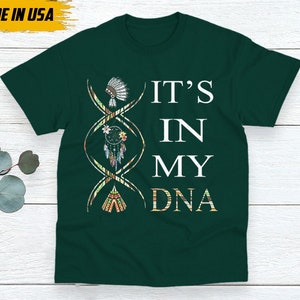 May include: Dark green t-shirt with the text "IT'S IN MY DNA" in white. The design includes a DNA strand with Native American-inspired graphics, including a headdress, dreamcatcher, and teepee. The words "MADE IN USA" are in the upper left corner.