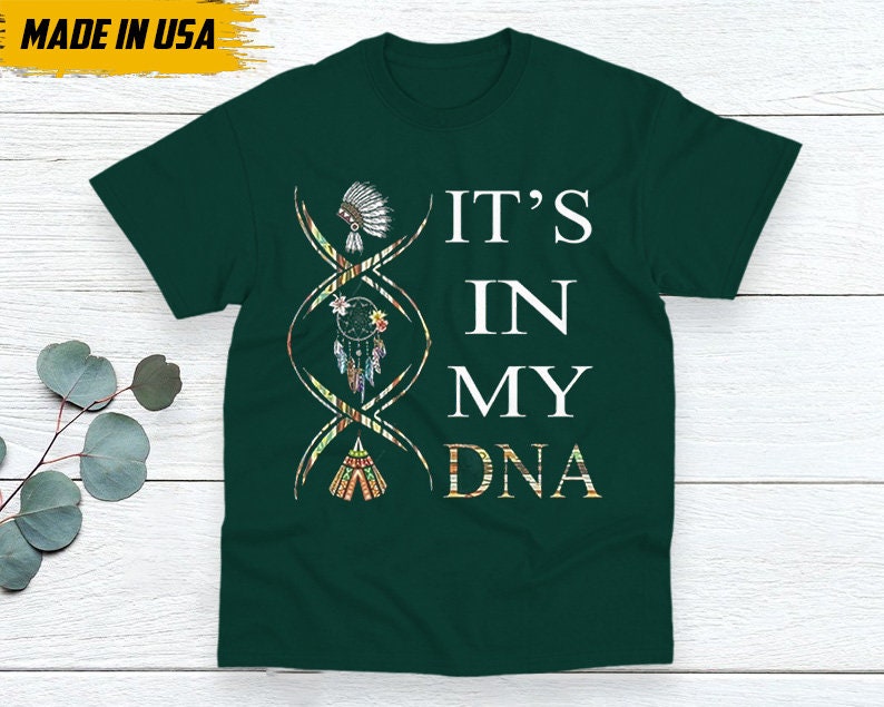 May include: Dark green t-shirt with the text "IT'S IN MY DNA" in white. The design includes a DNA strand with Native American-inspired graphics, including a headdress, dreamcatcher, and teepee. The words "MADE IN USA" are in the upper left corner.