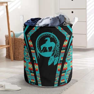 May include: A cylindrical laundry hamper with a Native American design. The hamper features a black background with a turquoise dreamcatcher design and a silhouette of a person on a horse. The sides have a turquoise, red, and yellow geometric pattern. White handles are attached to the top.
