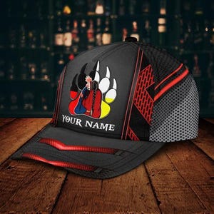 May include: Black baseball cap with a red and black design. The cap features a graphic of a person in traditional clothing inside a bear paw print. The text "YOUR NAME" is printed on the front.