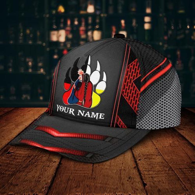 May include: Black baseball cap with a red and black design. The cap features a graphic of a person in traditional clothing inside a bear paw print. The text "YOUR NAME" is printed on the front.