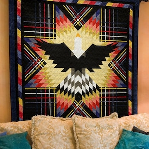 May include: A large, geometric quilt featuring an eagle design. The quilt has a black background with a central eagle motif in black, white, and gold. The design is framed by a navy blue border and accented with red, yellow, and white plaid patterns. The quilt is displayed on a wall.