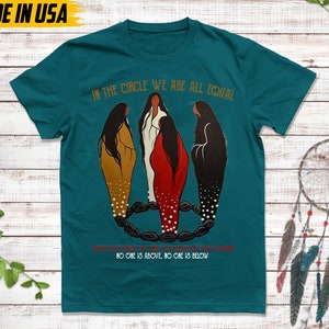 Puede incluir: Camiseta de color turquesa con el texto "IN THE CIRCLE WE ARE ALL EQUAL" y una ilustraci&oacute;n de tres figuras en marr&oacute;n, blanco y rojo. El texto "NO ONE IS ABOVE, NO ONE IS BELOW" tambi&eacute;n est&aacute; en la camiseta. "MADE IN USA" est&aacute; impreso en la parte superior izquierda.