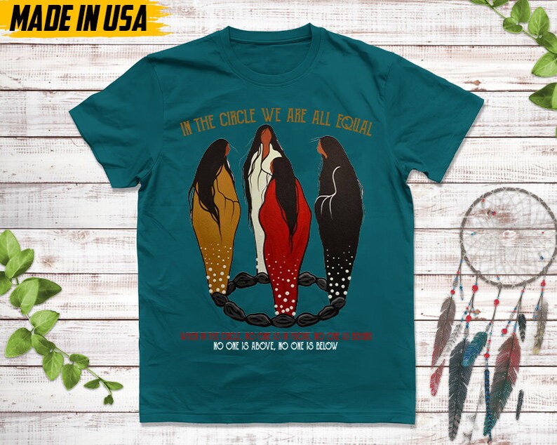 Puede incluir: Camiseta de color turquesa con el texto "IN THE CIRCLE WE ARE ALL EQUAL" y una ilustraci&oacute;n de tres figuras en marr&oacute;n, blanco y rojo. El texto "NO ONE IS ABOVE, NO ONE IS BELOW" tambi&eacute;n est&aacute; en la camiseta. "MADE IN USA" est&aacute; impreso en la parte superior izquierda.