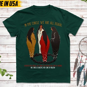 Puede incluir: Camiseta verde oscuro con el texto "IN THE CIRCLE WE ARE ALL EQUAL". La camiseta presenta una ilustraci&oacute;n de tres figuras con vestidos largos, con el texto "WHEN OF THE CIRCLE, NO ONE IS ABOVE, NO ONE IS BELOW". Las palabras "MADE IN USA" est&aacute;n en la parte superior.