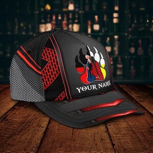 May include: Black baseball cap with a mesh back and red accents. The cap features a graphic of a Native American figure within a bear paw print, with the text "YOUR NAME" below the graphic. The cap is on a wooden surface.